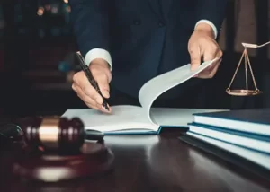 A person in a suit signs a document on a desk with a gavel, scales of justice, and law books.